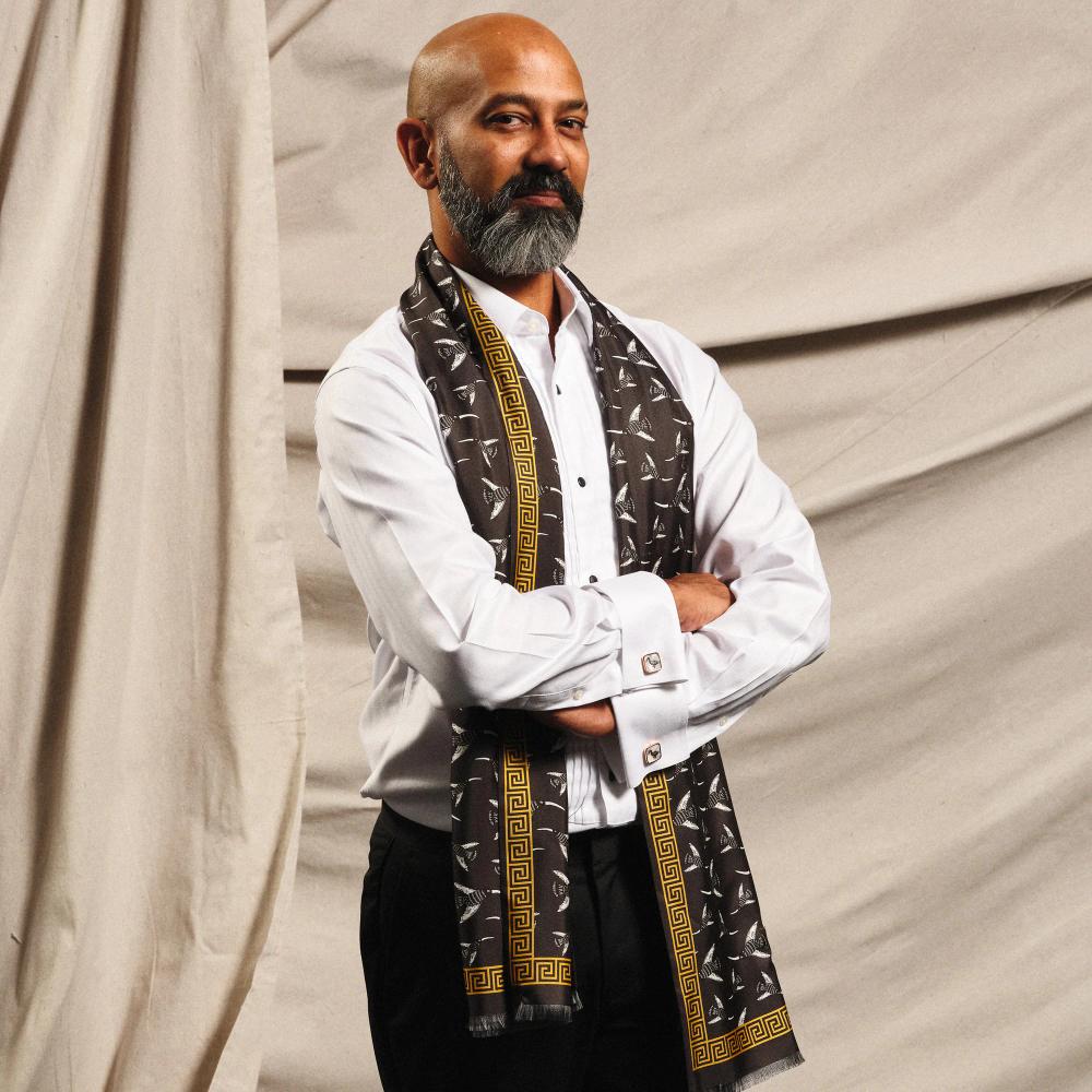 The artist Brendan Fernandes, a bald brown man with a dark beard, models his long scarf. Its gold border resembles a Greek-style spiral frieze. The interior pattern features illustrated birds with long beaks flying in different directions on a black ground.