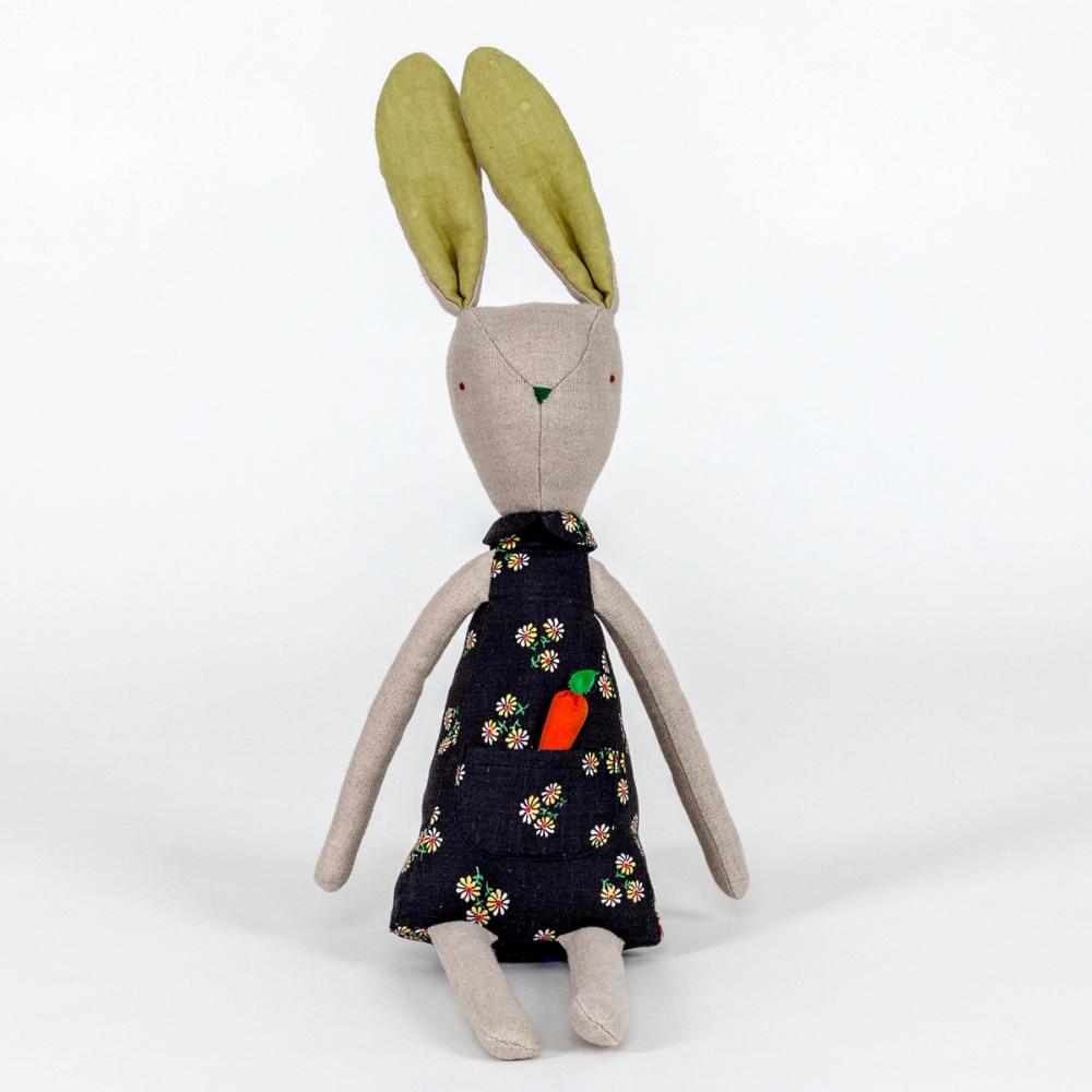 A photograph of a plush rabbit made of light beige woven fabric characterized to have a pointy nose, triangular shaped body, and long thin arms and legs. The body of the rabbit is made of a primarily blue patterned fabric and includes a pocket with a removable plush orange carrot.