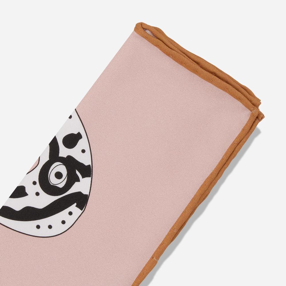 A detail of a folded rose pink silk pocket square with a warm, dark tan bordered seam. There is a glimpse of the printed bold black and white illustration of the Bata bird.