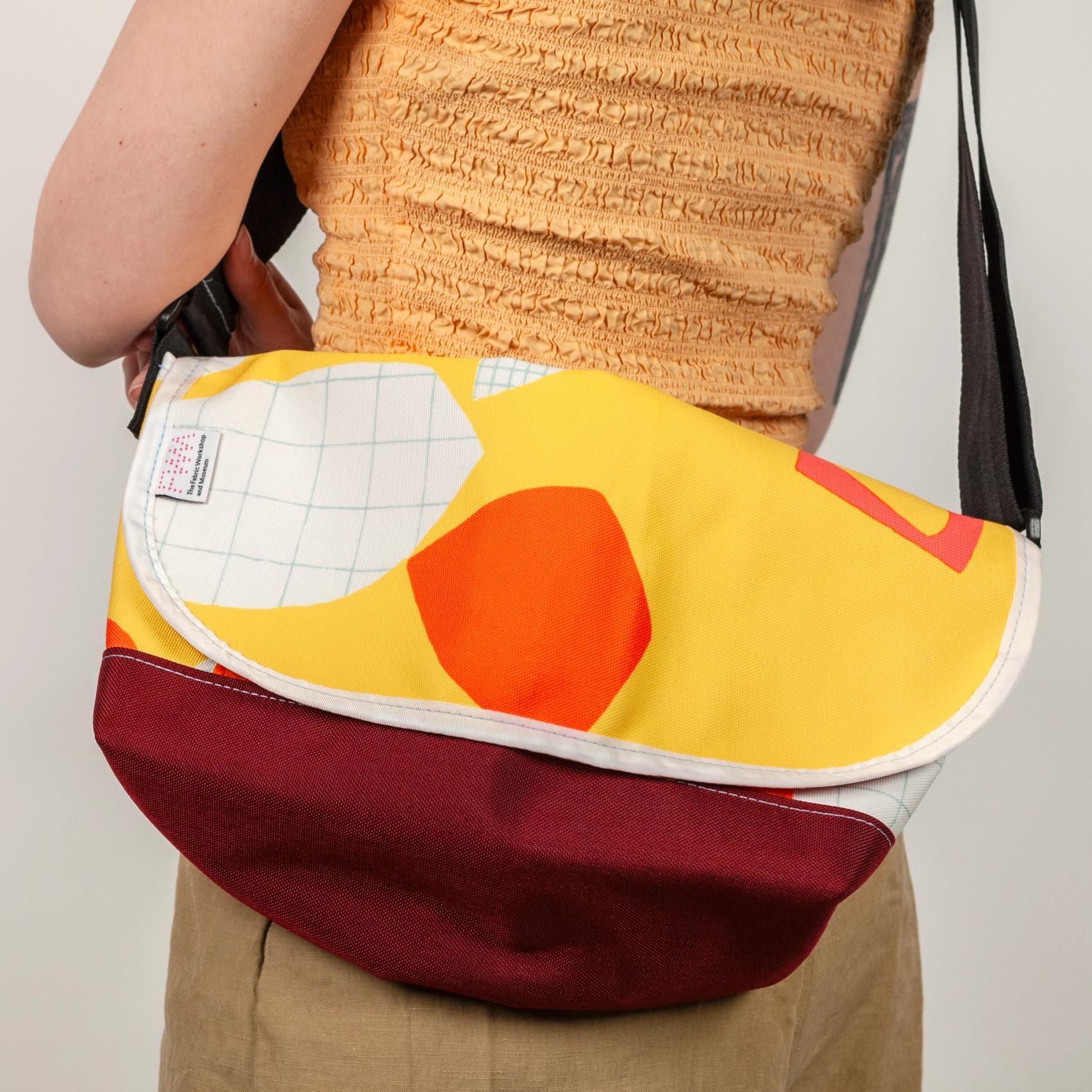 A model wearing a vibrant yellow bag with bold abstract shapes (bright orange and white with a light blue grid pattern) on her shoulder. The bag has a velcro top flap closure with a white seam along the flap. Along the white seam on left side of the bag is a white tag with a pink dotted logo reading "FWM" with "The Fabric Workshop and Museum" written underneath  The bag has a black strap and the base of the bag is solid maroon.