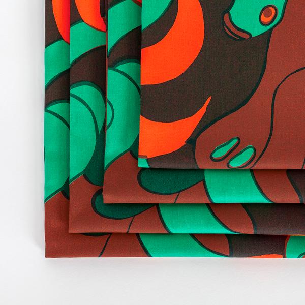 A detail photograph of a set of four square napkins made of fabric featuring a repeating textile pattern with stylized green snakes intertwined with abstract brown shapes and bright orange spirals on a dark brown background. The napkins are folded in squares atop one another.
