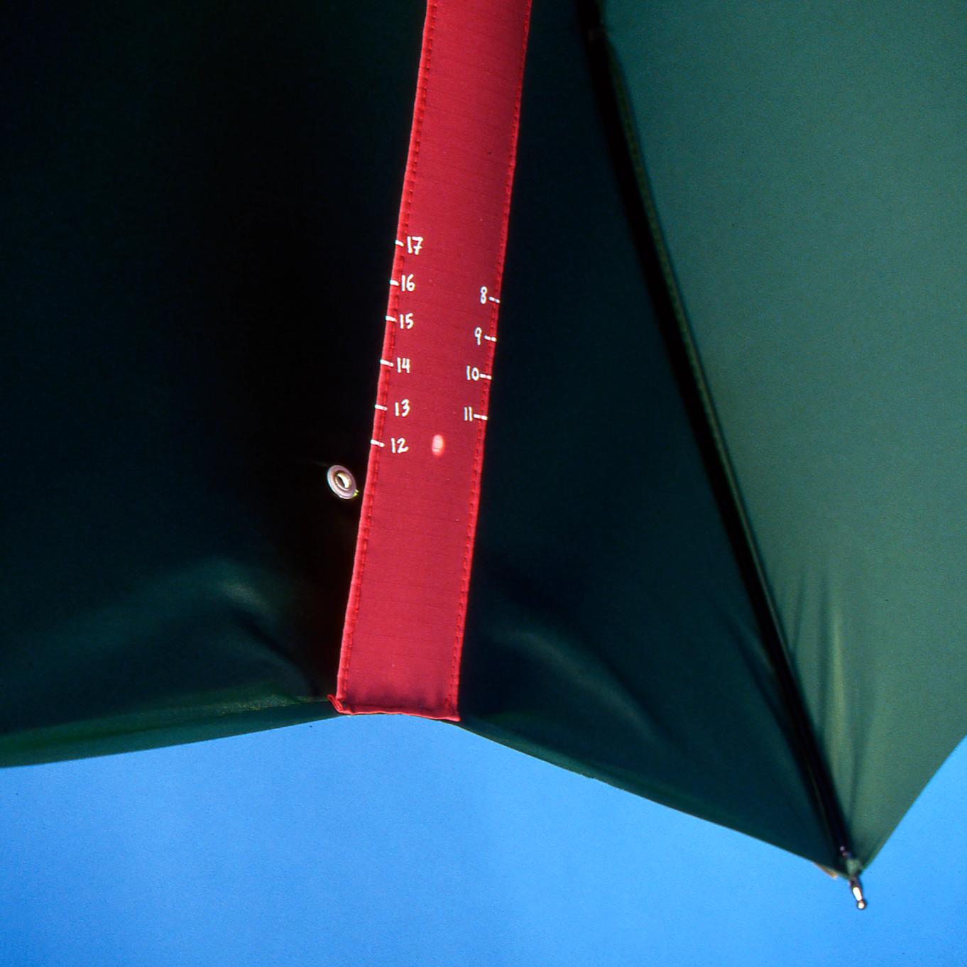 A detail photograph of the inside of a green umbrella with a wooden handle. There is a red strip of fabric sewn from the top to the bottom of the umbrella, suggesting to be used as a sun dial and marked with numbers 8 through 17. There is a dot of sunlight peeking through an eyelet on the umbrella, marking 12:00 on the sundial.