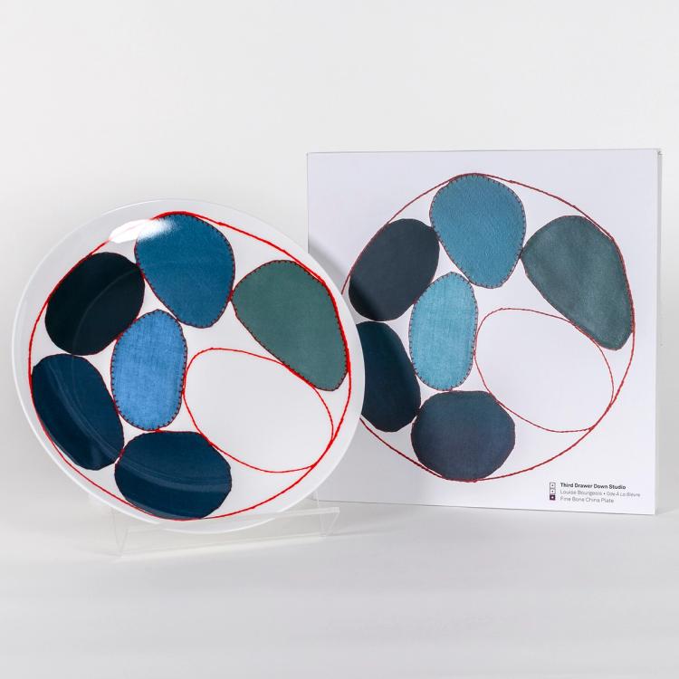 A photograph of a white plate with circles outlined in a red stitch design containing different fabric textures and shades of blue in the center. The circles are of varying sizes and shapes and they are encompassed in a larger red "stitched" circle bordering the outline of the plate. The plate is standing upright beside a white box featuring a photograph of the plate.