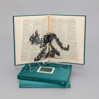 A photograph of a book spread atop two stacked green books that read "William Kentridge Lexicon" on the spine. The book spread shows a frenetic drawing in black ink over tiny, dense text. The drawing resembles the profile of a cat with its head facing left and its tail curled slightly upward on the right page.