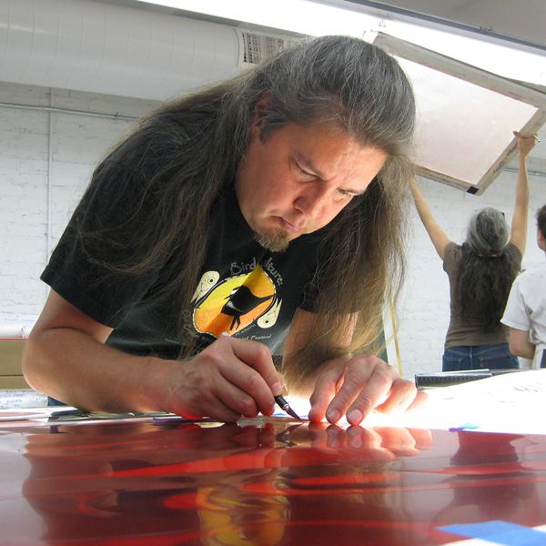 The artist Tommy Joseph, a Native American man with long greying dark hair, leans over a light table with a drawing tool in his right hand. A person in the background holds a screen up to the florescent light.