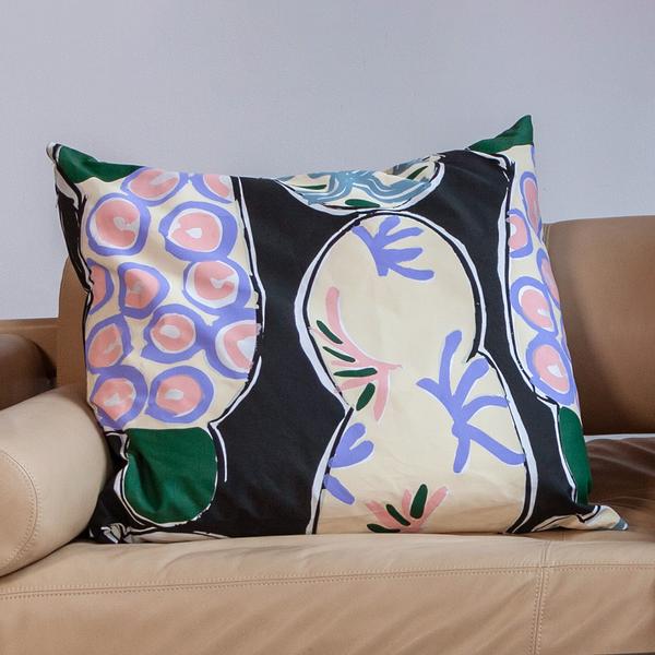 A square pillow leans against a beige couch. The pillow is a screenprinted design by Betty Woodman. The background of the design is black, with cool and warm pastel colors, and loosely drawn, soft abstract shapes mimicking ceramic platters.