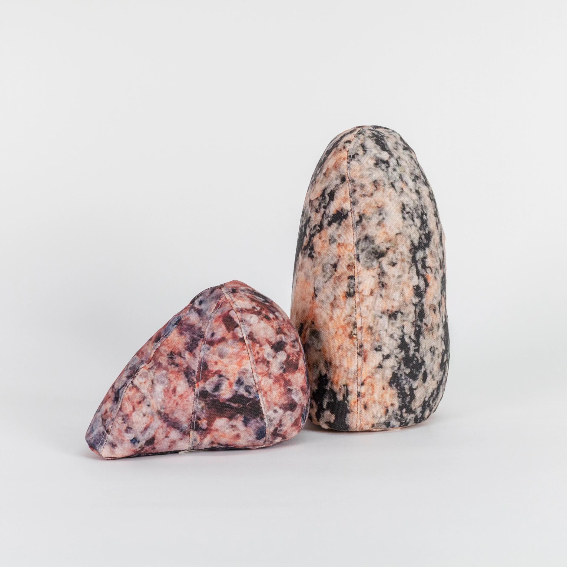 A photograph of two small soft sculptures covered with patterns that resemble polished stone of different hues. One sculpture is short and triangular where the other one is tall like an obelisk with a rounded top.