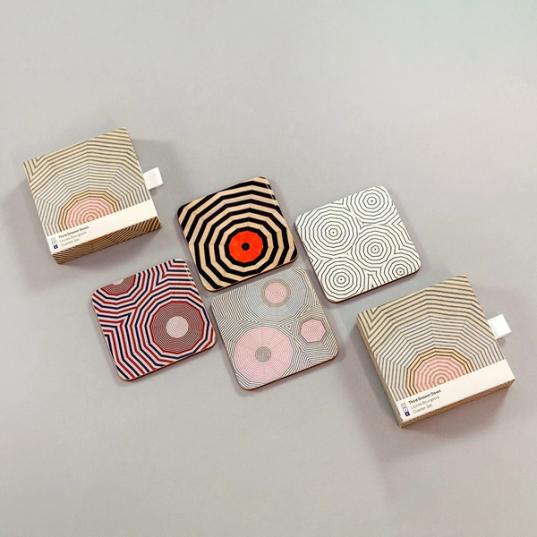 An image of the same set of four coasters in a similar arrangement, but from a further away perspective and showing the coaster packaging as it framed the coasters on the top left and right bottom. The packaging is a square box featuring another geometric design by Louise Bourgeois with a small text label at the bottom which reads "Third Drawer Down, Louise Bourgeois, Coaster Set"
