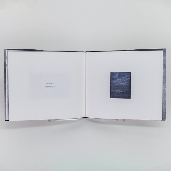 A photograph of an open book. On the left is a mostly blank page except for four short rows of text. On the right is a small colored pencil landscape drawing on black paper.