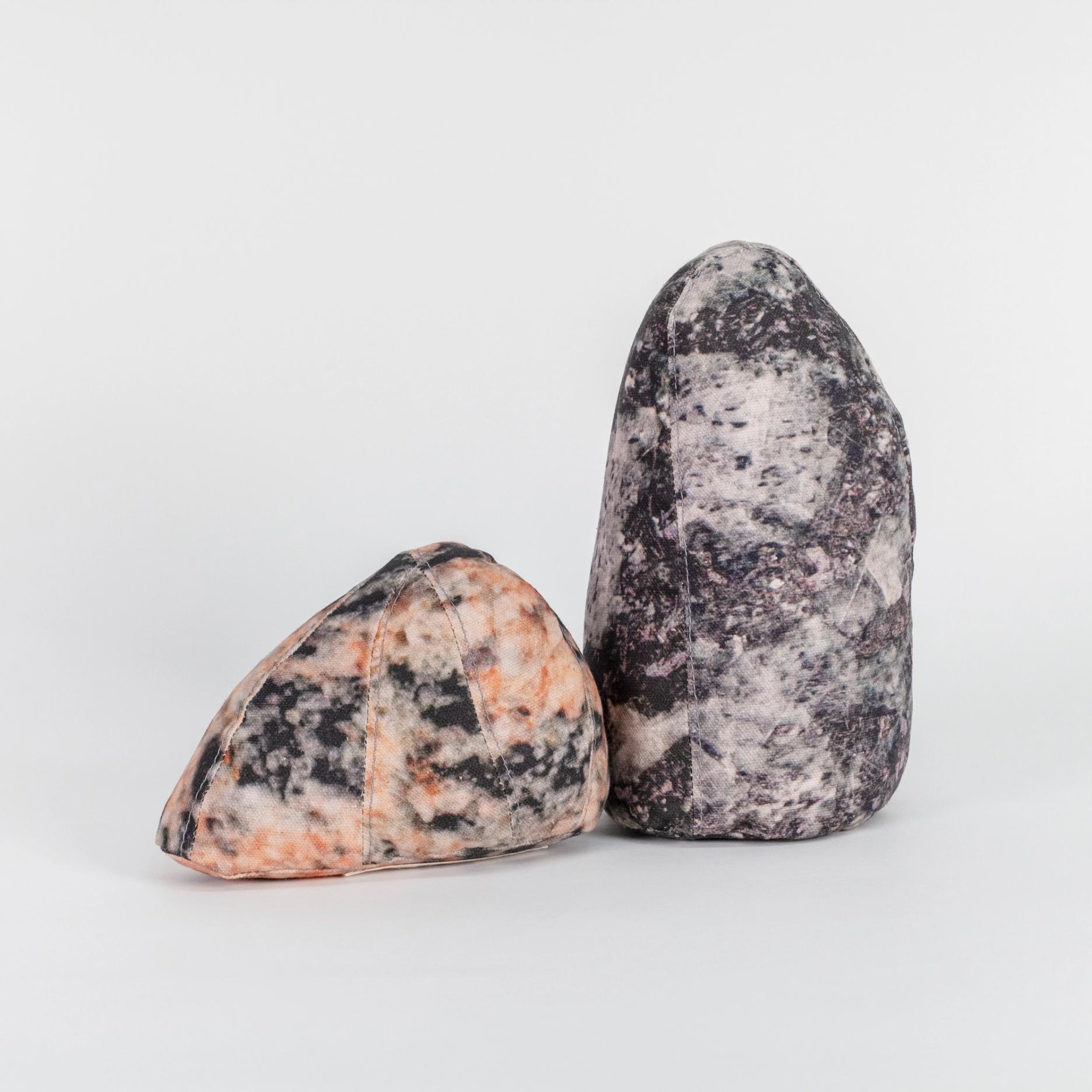A photograph of two small soft sculptures covered with patterns that resemble polished stone of different hues. One sculpture is short and triangular where the other one is tall like an obelisk with a rounded top.