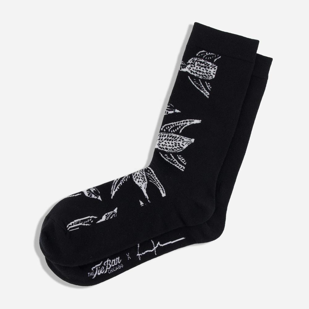 A pair of black knit dress socks with a pattern of white illustrated Shomoro birds flying in different directions. The artist's signature and "The Tie Bar" logo is featured on the bottom of the socks.