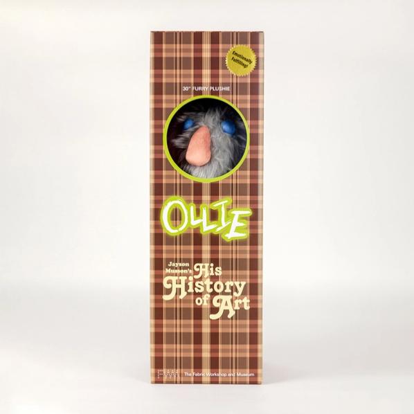 A photo of a tall brown plaid box featuring a circular viewing window that reveals a gray stuffed animal inside. Above the viewing window it says "30 inch furry plushie" and includes a gold sticker that says "emotionally fulfilling!" Below the window it reads "Ollie" and "Jayson Musson's His History of Art"