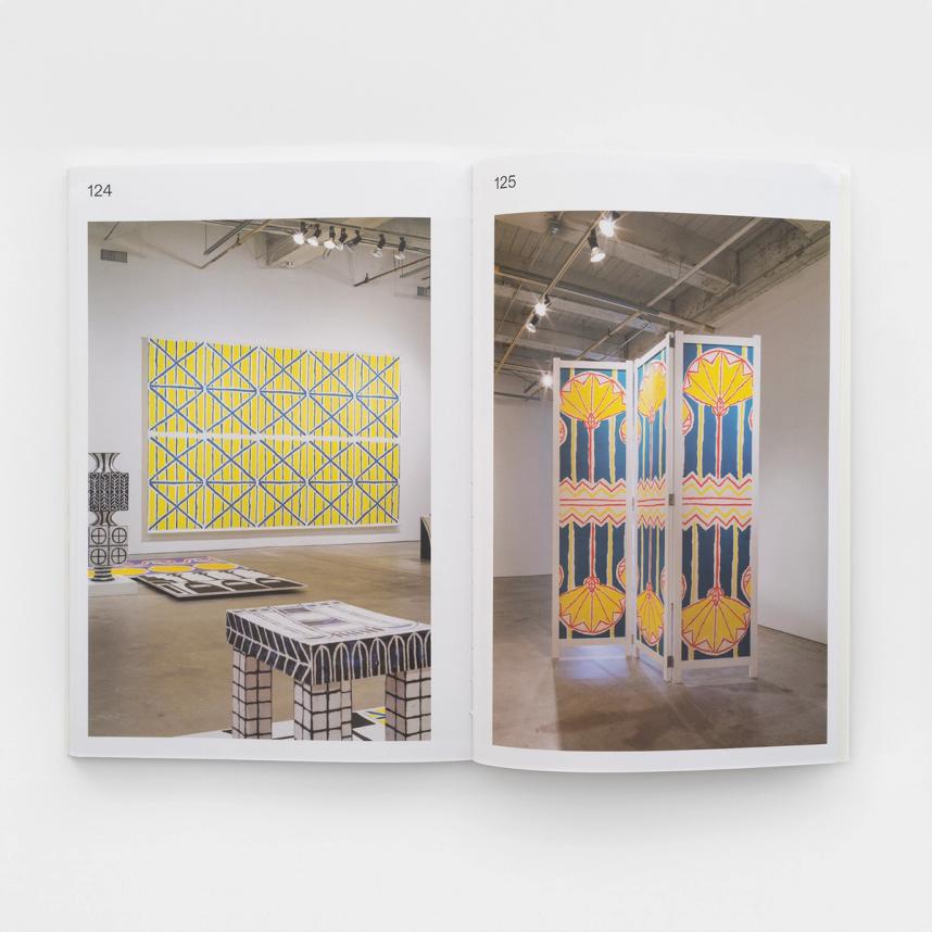An image of a book spread open to two large photographs, one on each page. On the left is an exhibition view of a large, geometric, abstract reapeat pattern in yellow, teal, and white. It is the same pattern that is on the cover of the book. It is a screenprint and is hanging on the wall like a large painting. In the foreground there are three sculptures decorated with geometric patterns. Two of the sculptures are made of ceramic and are black and white. The third sculpture is a flat panel on the ground with two geometric patterns, one black and white and the other is yellow, purple, and red. On the right is an exhibition view image of a room divider, decorated with a yellow, red, and teal repeat geometric pattern.