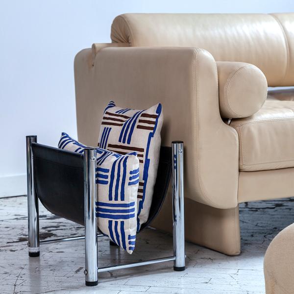 Two pillows rest inside a low rack beside a couch. The rectangular pillows feature cream-colored fabric with a minimal weave-like pattern of blue and brown stripes in groups of three.