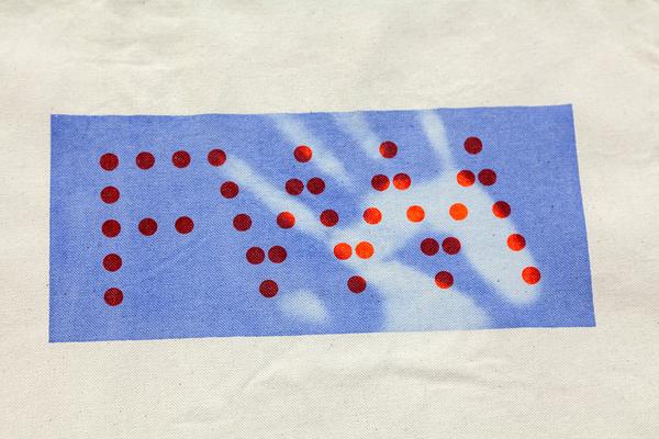 A detail photo of a light tan canvas tote bag. On the bag, there is a screenprinted light blue rectangle (landscape). Inside the light blue rectangle are the letters "FWM" and the letters are made up of bold, dark red dots. There is a faded spot on the rectangle in the shape of a hand from the hand model pressing their hand on the tote on top of the screenprinted light blue rectangle. The light blue is now a pale, almost white-blue. The dark red dots are now bright orange.
