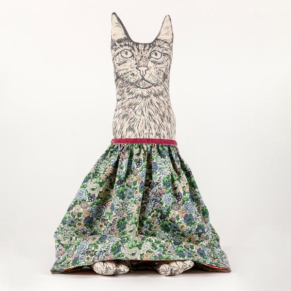 A photograph of a reversible flip doll. In this instance, a plush cat with drawn details stands upright with a skirt of florals screenprinted in various greens with some blues and earth tones. The cat has little feet peeking out from beneath the skirt's hem.