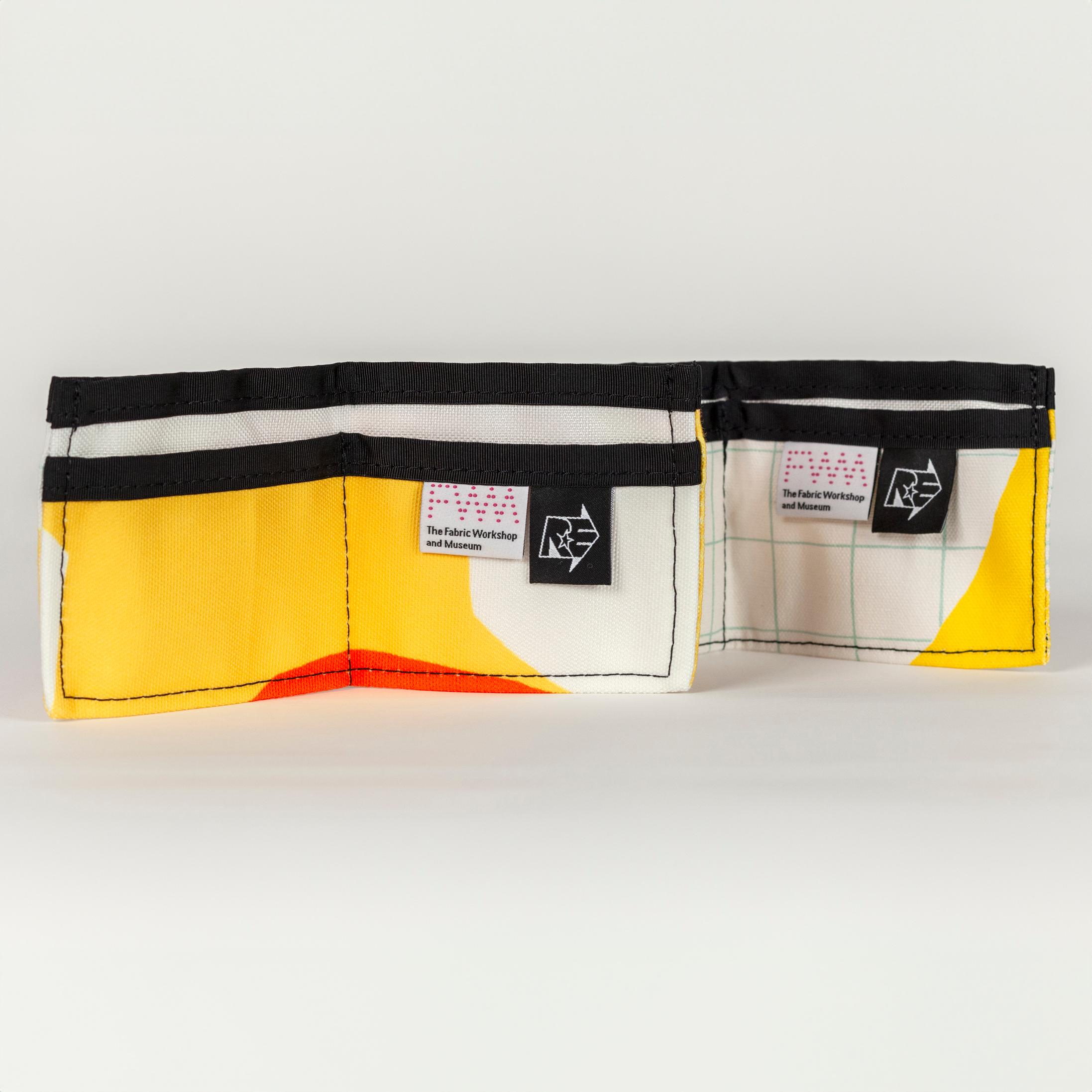 Two open vibrant yellow wallets (opens into a wide rectangle with one large pocket in the back and two smaller pockets in front) with bold abstract shapes (bright orange, white, and white with a light blue grid pattern). There is a solid black seam lining the top of the wallet. There are two tags on the right pocket. The tag on the left is white with a pink dotted logo reading "FWM" with "The Fabric Workshop and Museum" written underneath. The tag on the right is black with an arrow. Inside the arrow are the letters "RE"