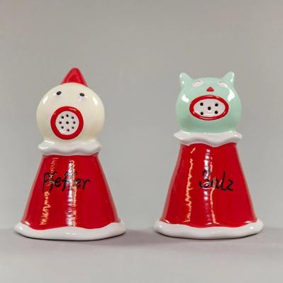 A photograph of a pair of salt and pepper shakers resembling cartoon-like characters against a light gray background. On the left, a triangular-shaped pepper shaker takes the form of a jester. A pointed red hat sits atop its spherical cream head. It has two black dots for eyes and red lips encircling nine tiny pour spouts. Its head sits above a wavy white collar and a red dress reading "Pfeffer" with white trim. The salt shaker to the right resembles a cat with the same red dress with white trim, reading "Salz". The cat's sea-foam green head is round with two nubby ears, a small red triangle for a nose, and an ovular red mouth encircling five tiny holes for sprinkling salt.