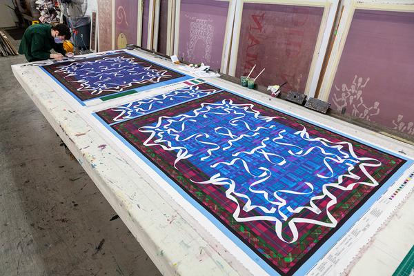 A photograph showing two long textile designs with a dark red and green border and a blue center. A mirrored pattern with white ribbons is printed over the blue and extends over the border into the red. An artist works on the far left of the photograph at the end of the table.