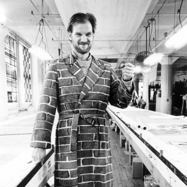 A black and white photograph of a white man posing for the camera with his left fist partially raised near at shoulder-height. He is wearing a long jacket featuring a brick pattern. He is standing in a well lit studio with long printmaking tables extending behind him from each of his sides.