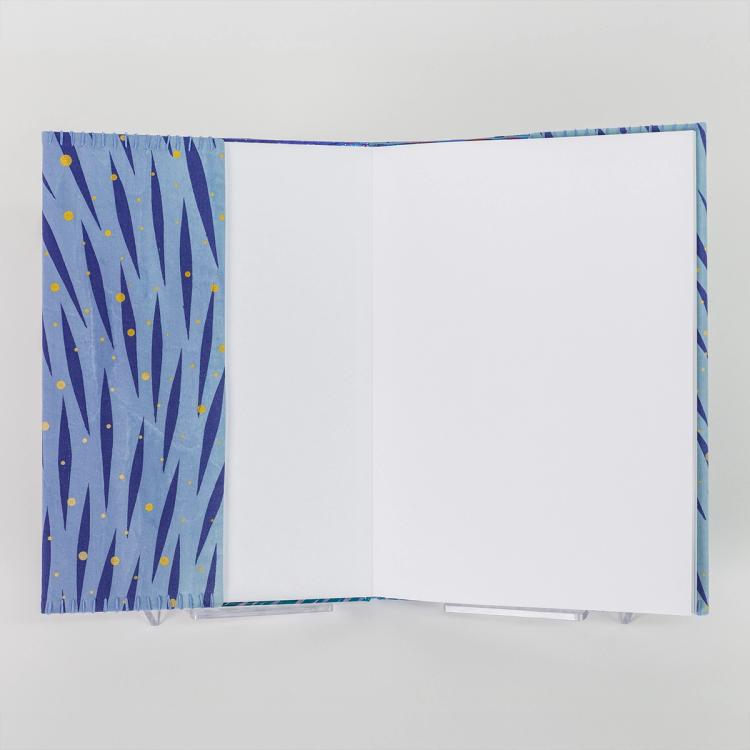 A photo of a hardback book open to the first blank pages featuring the slip of the fabric book cover with a hand-stitched seam and a vibrant pattern of diagonal dark blue brush-like streaks and scattered yellow dots on a light blue background.