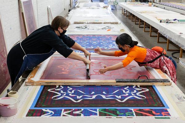 A photograph of two artists leaning over a long table to screenprint together. The pattern they are printing is made up of dark green, red, and blue with white lines.