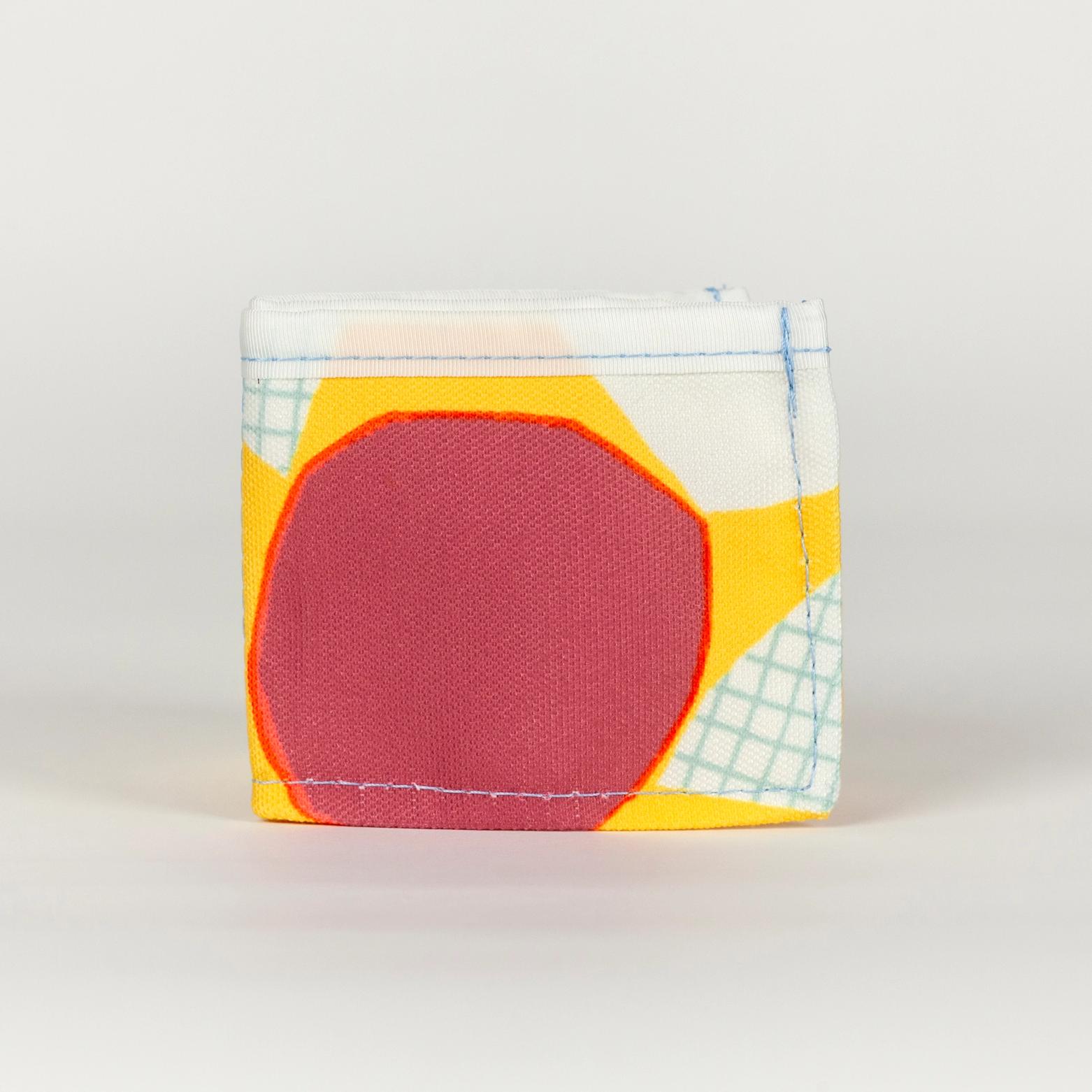 A vibrant yellow wallet folded in half (from being a wide rectangle into a square) with bold abstract shapes (bright purple, white, and white with a light blue grid pattern). There is a solid white seam lining the top of the wallet.