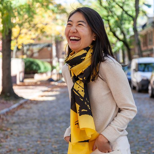 A laughing woman wearing a silk scarf stands along a city street on an autumn day. Her scarf is yellow and features bold, painterly gestures with ink drips and splatters that follow the curving motion of a brush.