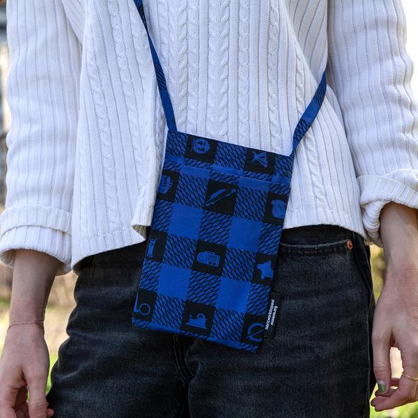 A model wears a small flat bag at her hip. The bag features a blue and black plaid pattern with icons of objects in the black squares.