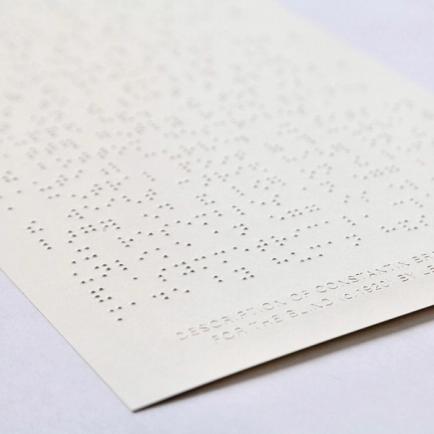 A detail shot of the item—a cream colored piece of paper with writing in braille—sitting on a white backdrop.