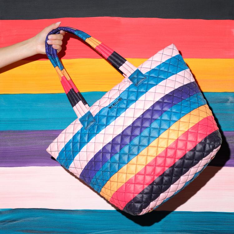 A photograph of a model holding out a fabric quilted tote bag featuring painted horizontal stripes of alternating light pink, purple, blue, ochre, red, and black. The bag is in front of a painting with the same horizontal striped pattern.