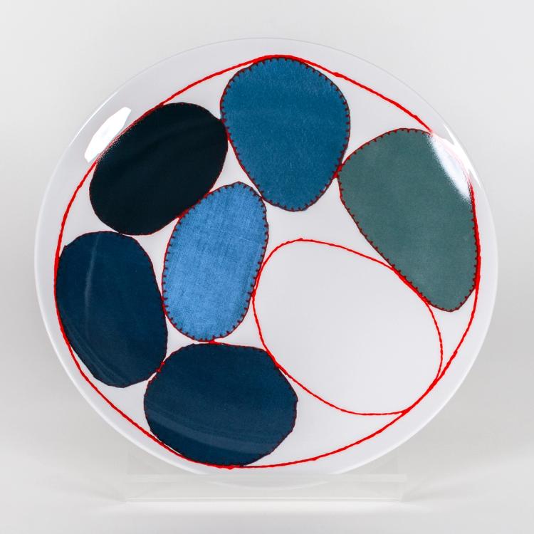 A photograph of a white plate with circles outlined in a red stitch design containing different fabric textures and shades of blue in the center. The circles are of varying sizes and shapes and they are encompassed in a larger red "stitched" circle bordering the outline of the plate.