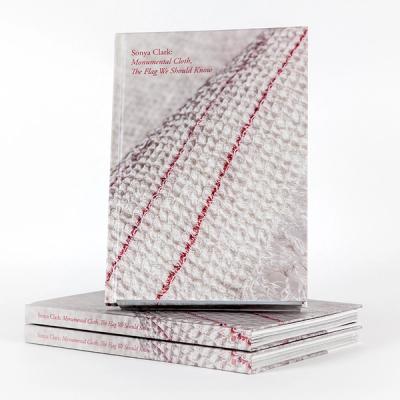 A photograph of a book propped upright on top of a pair of laterally stacked copies of the same book. The cover of the book features a cream-colored woven textile with two thin red stripes that stretch from corner to corner. The text on the top-left corner reads, "Sonya Clark: Monumental Cloth, The Flag We Should Know."