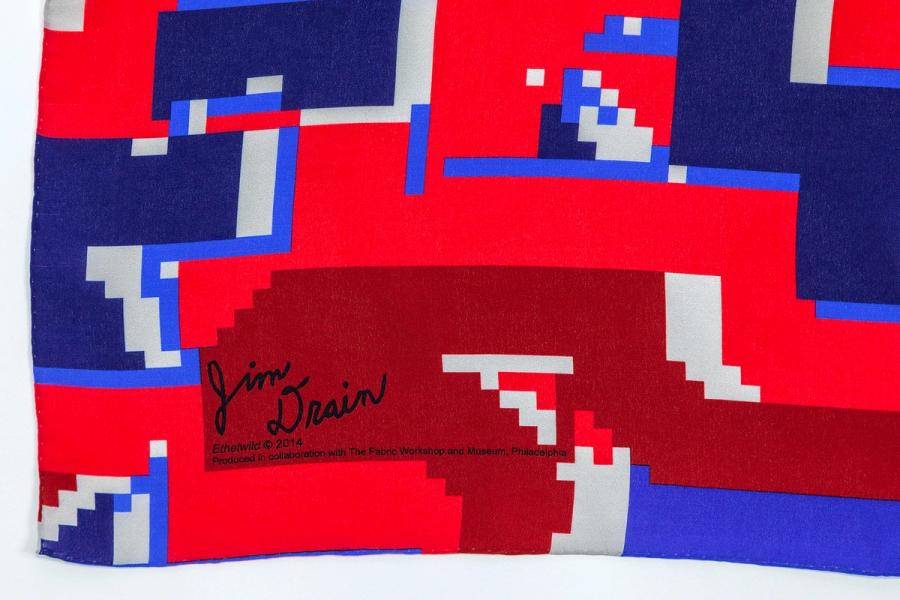 A corner detail of a scarf printed in pixelated blues, reds, and white shows the signature of the artist Jim Drain. 