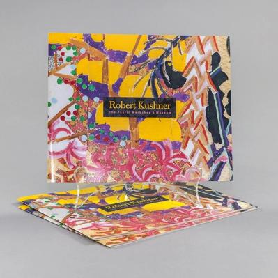 A thin paperback book, wider than it is tall, stands upright atop two other copies. Mostly yellow with some areas of white tan and black, the book features an abstract painting consisting of many repeated brushstrokes in red, purple, and tan. At its center is the title of the book in gold type on a black rectangle : Robert Kushner, The Fabric Workshop and Museum.