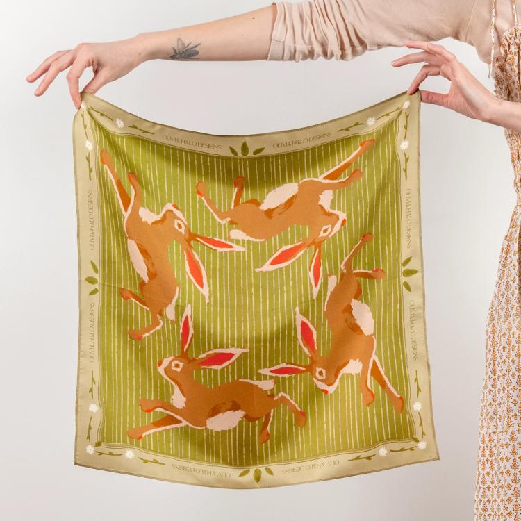 A photograph of a person holding up adigitally printed green silk scarf featuring brown rabbits running on each side of the scarf. Along the border are flowers and leaves and text that reads "Olivia Halo Designs"