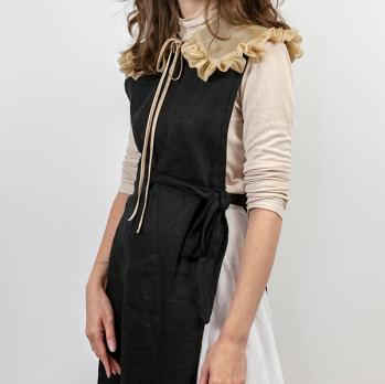 The collar in tea is being worn by a woman with brown hair who is standing against a white background. Underneath the collar, she is wearing a black sleeveless apron tied at the waist, on top of a white long-sleeved turtleneck shirt and white skirt. The shirt is pushed up to her elbows.