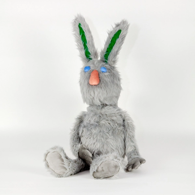 A gray plush stuffed rabbit sits upright. It has round blue eyes, an oblong peach noes, and long green shapes on the interior of its ears.
