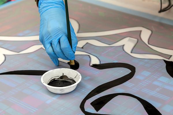A photographic detail of a hand in a blue glove painting with black ink into organic shaped lines. A shallow cup of black ink sits on the surface of the design quite close to the hand.