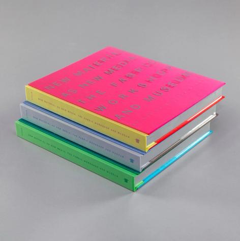 A photograph of a stack of three slightly offset books of the same size but with different color spines (green, gray, and yellow). The top book with a yellow spine is magenta on its cover. The book reads "New Material as New Media: The Fabric Workshop and Museum."
