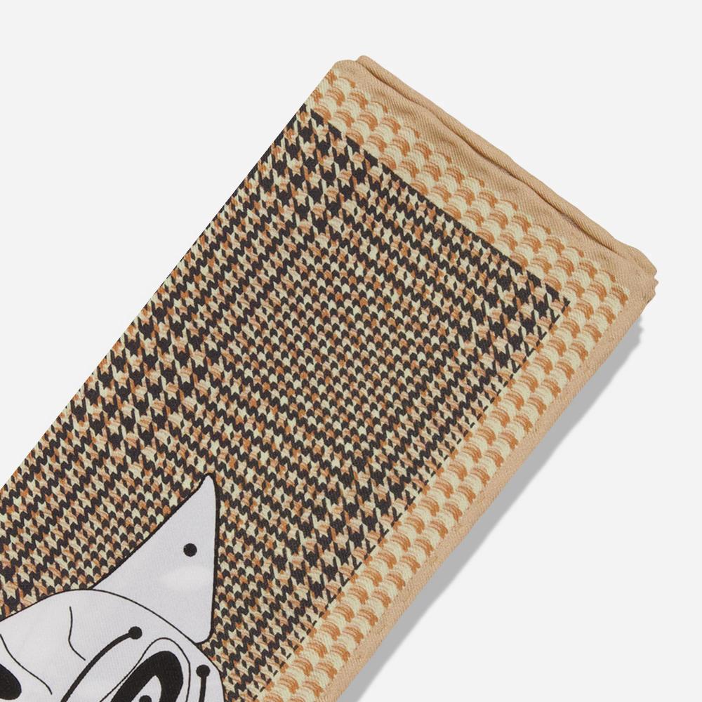 A folded pocket square with a detail of the corner. Its tan border resembles a woven texture while the rest shows a black and tan houndstooth pattern. There is a black line illustration of a house sparrow’s peak emerging from the bottom of the textile.