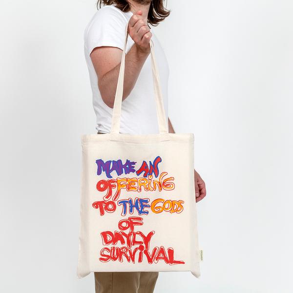A model holds out a cream-colored tote that reads in a hand-drawn message, "Make An Offering to the Gods of Daily Survival." The word daily is spelled d-a-y-l-y. Half the words appear in red while others are colored in with purple, pink, burnt yellow, and blue.
