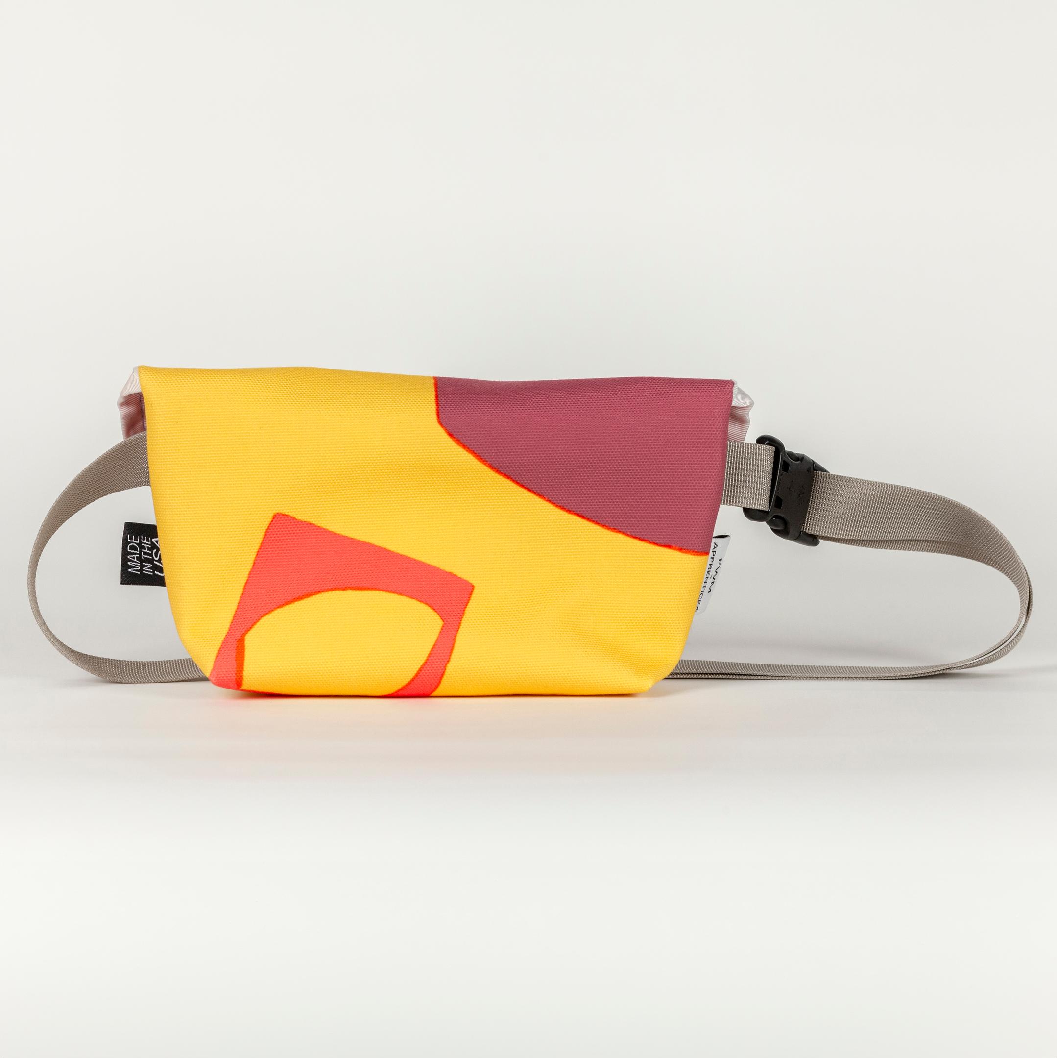 The back of a short, wide, vibrant yellow bag with bold abstract shapes (bright orange and purple). The bag has a shiny gray strap and the viewer can see one tag on the left side of the bag. The tag is black with text reading "MADE IN THE USA"