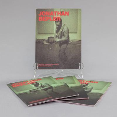 A photo of a book propped up and in the foreground more of the same book spread out as a display. The cover of the book features a grainy, green monotone photo of an artist playing guitar in a mostly empty room. There are two large acoustic panels on the wall.
