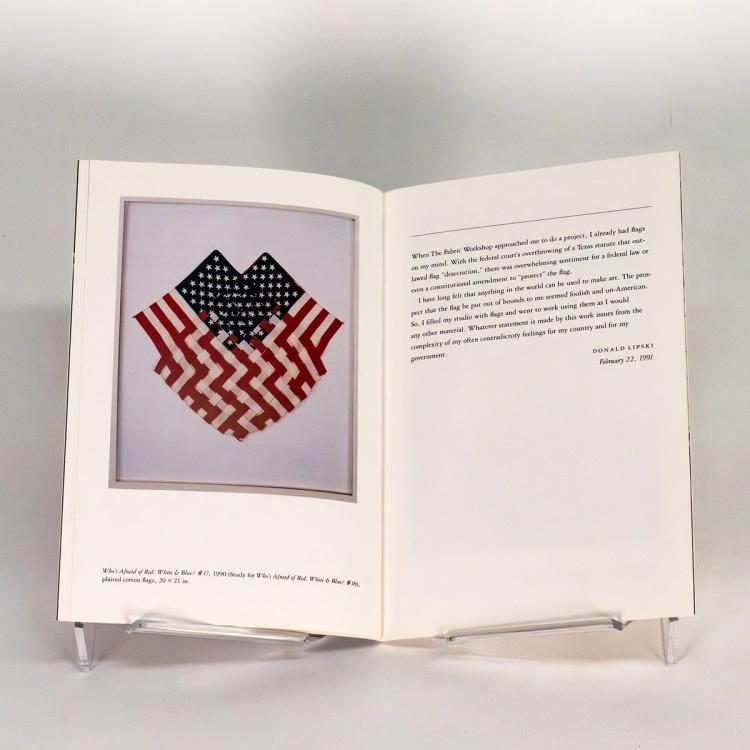 A photo of an open spread of a book. On the left is an image of two american flags woven together by the red and white stripes and displayed within a white frame. On the right is a description by the artist.