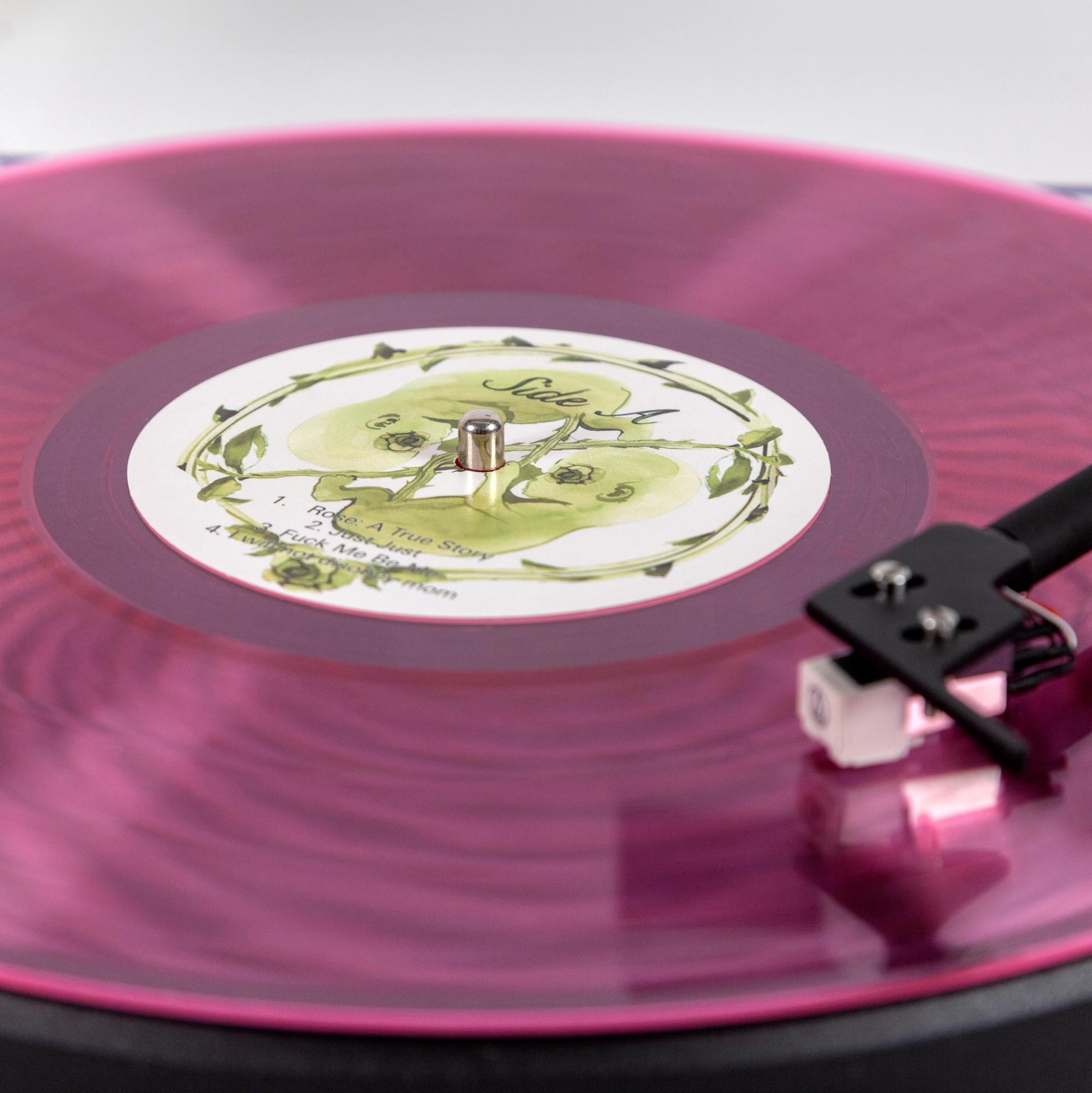 A detailed photo of a see-through pink vinyl record playing on a turntable. The center sticker reads "Side A" and features a motif of green hand-illustrated twin fetuses.