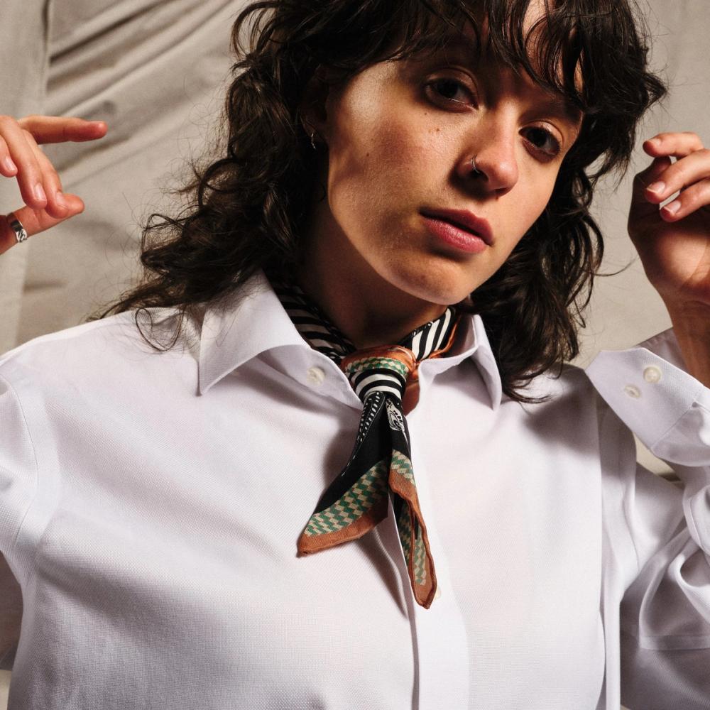 A model wears a small scarf tied around her neck. Each end features stripes of tan, followed by a houndstooth pattern of green and cream, followed by a broader black border over which illustrated birds are printed. Around the neck loop are black and white stripes.