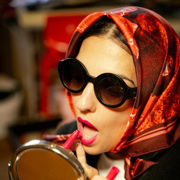 A woman models a red scarf with a mottled texture while applying lipstick in front of a handheld mirror. She wears sunglasses and the scarf over her head, her hair peeking out at the top.