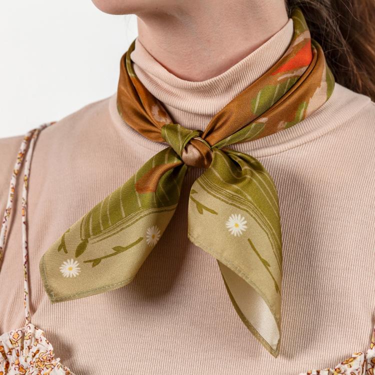 A photograph of a person wearing a green, brown, and pink digitally printed silk scarf tied around their neck. On the border of the scarf are white flowers and green stems and leaves.