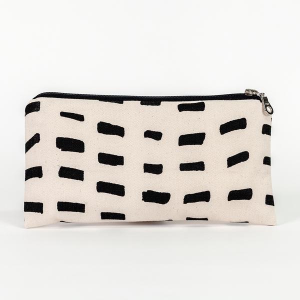 A canvas pouch, longer than it is tall, sits upright with a zipper resting on the top-right. Its design features a pattern of black dashes over a cream-colored ground.